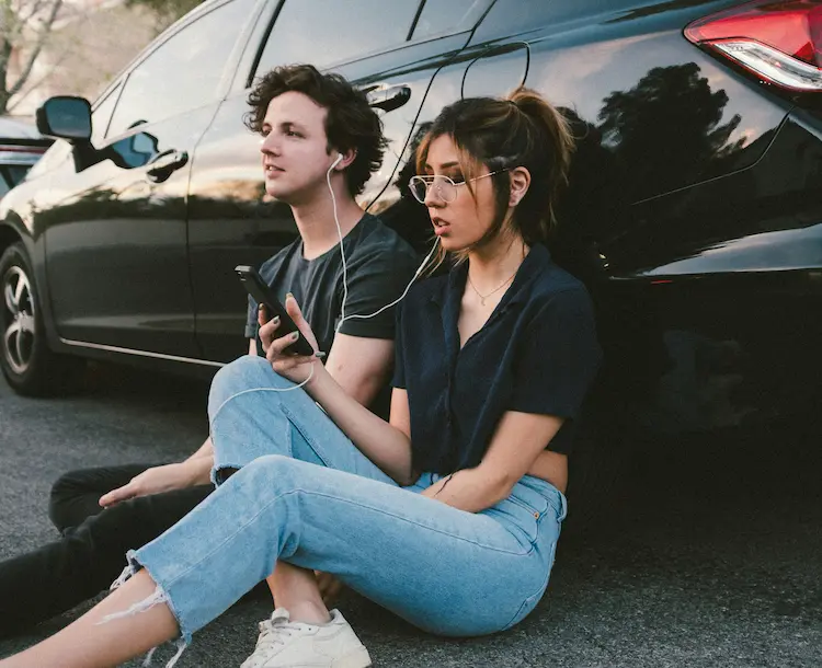A girl and a boy, sitting side by side, against a car, sharing a pair of earphones, listening to music