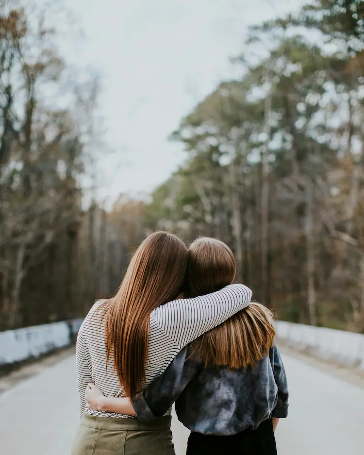 Two girls side hugging in the woods, showing their back sides.