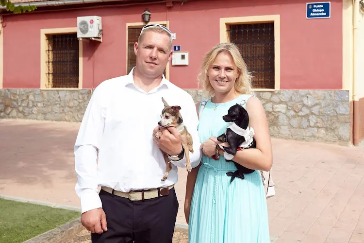 A blonde man in a white shirt on the left, holding a brown pup in his hands. A blonde woman, in a blue dress, on the right, holding a black pup in her hands.